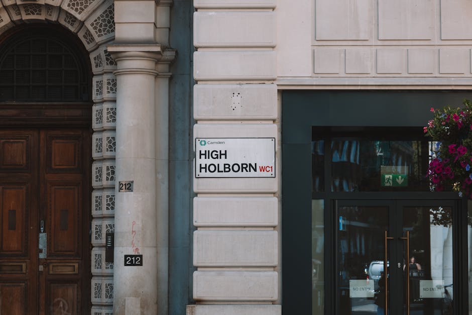 The image depicts the exterior of a building with a white stone facade, featuring an arched doorway with intricate detailing around the frame on the left side, and modern glass doors with metal handle